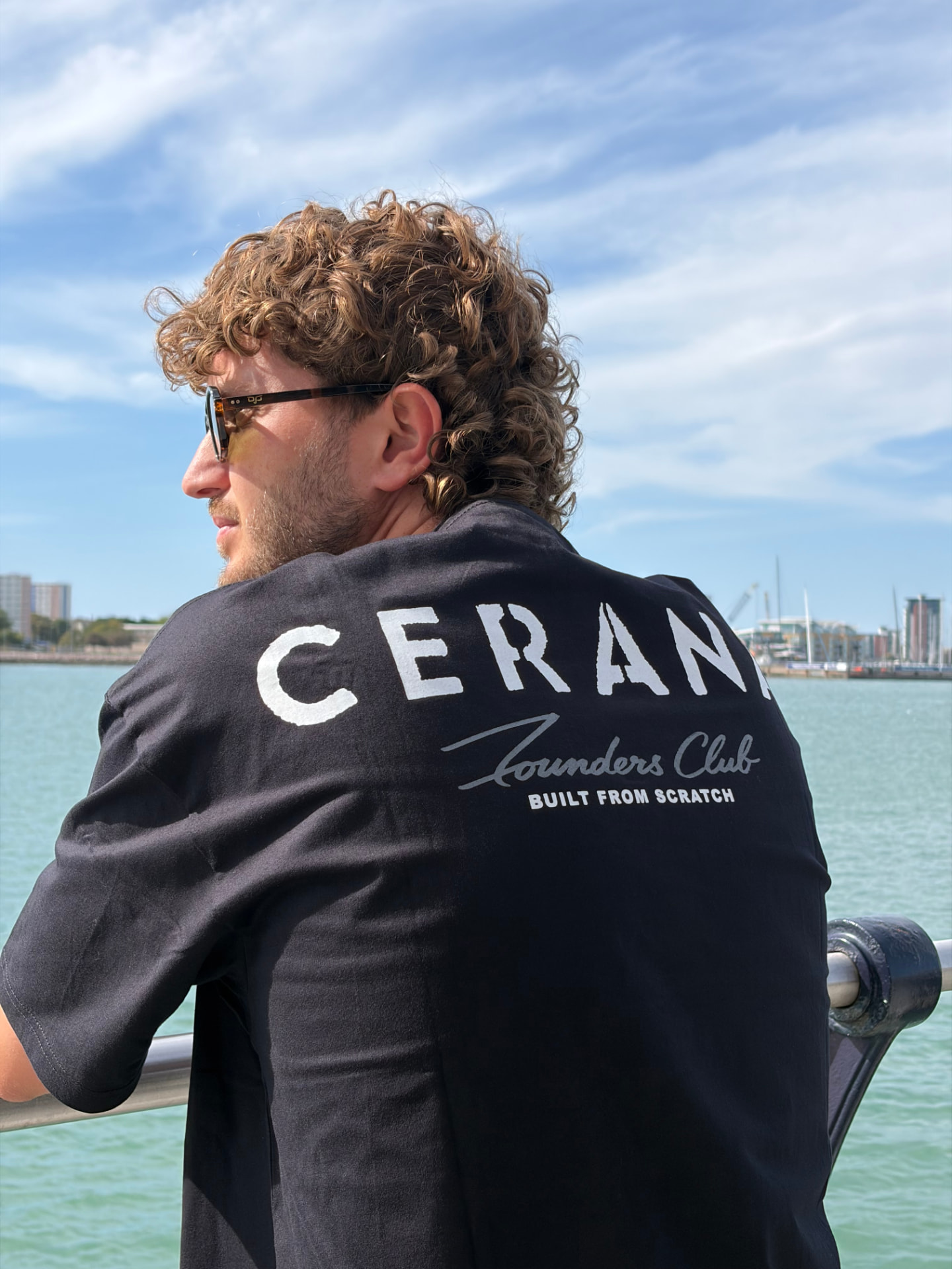 Cerana founder in a black Founders Club tee by the sea, back print, campaign tile.